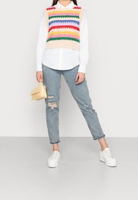 Persona che indossa un gilet di maglia a righe colorate sopra una camicia bianca, jeans blu strappati, sneakers bianche e tiene in mano una piccola borsa beige.