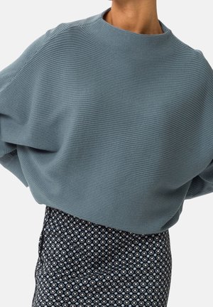 Kobieta ubrana w luźny, prążkowany, jasnoniebieski sweter w połączeniu z ciemną, wysoką spódnicą z wzorem na jednolitym tle.