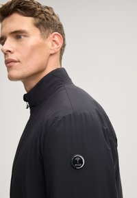 Giacca nera con zip e colletto alto, realizzata in tessuto liscio. Presenta una toppa rotonda con logo sulla manica sinistra. Fit aderente.