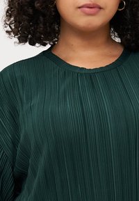 Άτομο με σγουρά μαλλιά φοράει μια υφασμάτινη, σκούρα πράσινη μπλούζα με πτυχώσεις, εστιάζοντας στη γραμμή του λαιμού και την άνω περιοχή του στήθους. Είναι εμφανές ότι έχει λείο δέρμα και ουδέτερο μακιγιάζ.