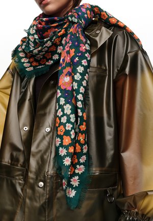 Mujer vistiendo una bufanda floral verde oscuro con flores naranjas, blancas y moradas sobre un impermeable verde oliva brillante con botones de presión.