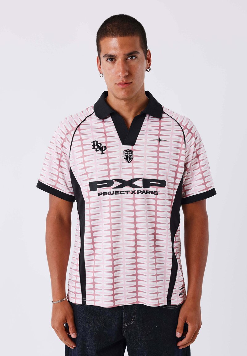 Homme portant un polo à manches courtes motif rose et blanc avec col noir et logo « Project X Paris », debout devant un fond uni.