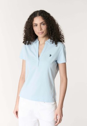 Donna con capelli ricci e scuri che indossa una polo a maniche corte azzurro chiaro e pantaloni bianchi, in piedi davanti a uno sfondo semplice.