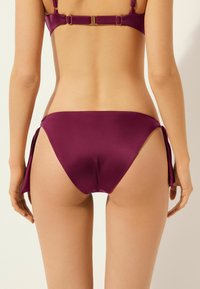Bikini bottom i djupt lila satin med sidband. Slät textur, låg midja och måttlig täckning bak.