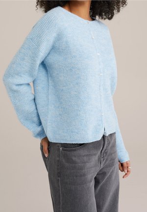 Femme portant un cardigan boutonné duveteux bleu clair et un jean taille haute gris foncé, la main droite dans la poche.