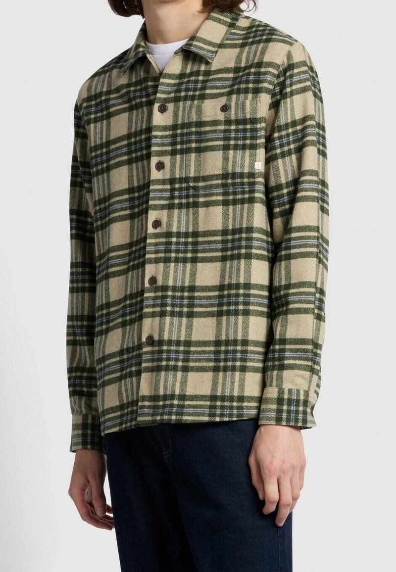 Personne portant une chemise à carreaux beige et verte boutonnée sur un sous-vêtement blanc avec un pantalon bleu foncé, debout devant un fond uni.