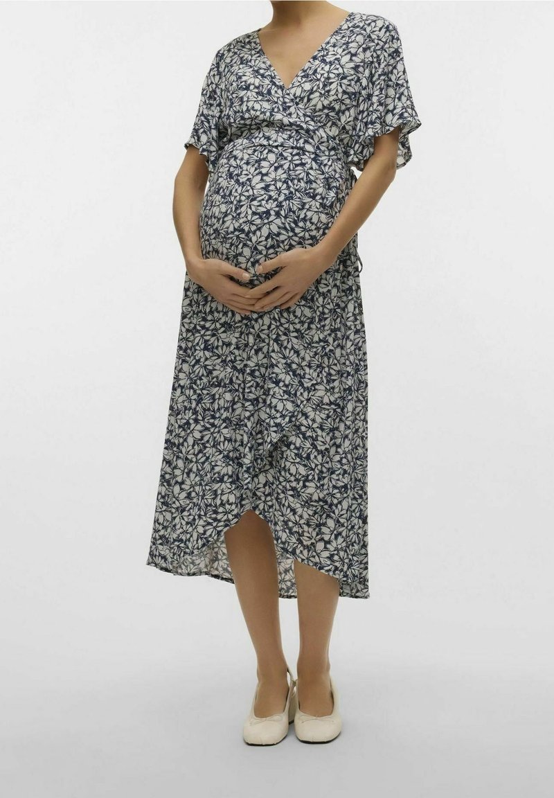 Femme enceinte en robe portefeuille fleurie bleu marine et blanche, tenant son ventre, portant des chaussures plates crème sur un fond uni.