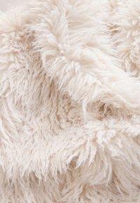 Strukturiertes, flauschiges Gewebe in einem elfenbeinfarbenen Farbton mit langen, weichen Fasern, die eine warme und einladende Oberfläche schaffen. Keine deutlichen Muster oder Akzente.