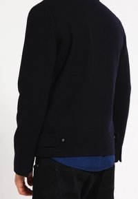 Schwarze Wolljacke mit hohem Kragen, glatter Textur und Metalldetails an den Gesäßtaschen, getragen über einem blauen Hemd.