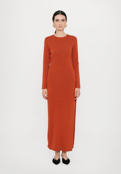 Robe ajustée à manches longues en tricot couleur orange brûlé, atteignant le mi-mollet. Elle présente un col rond et des fentes latérales, associée à des chaussures noires à talons pointus.