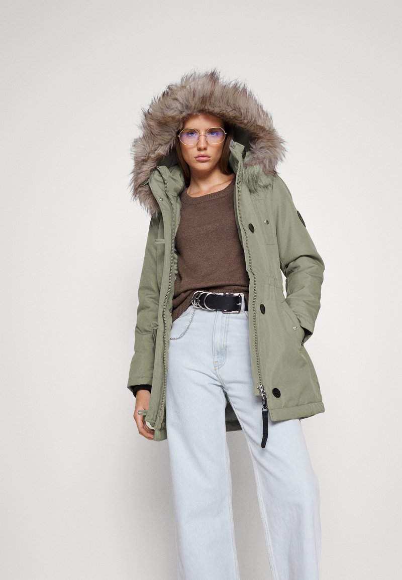 Parka olive avec une capuche doublée de fourrure, des boutons sur le devant et des détails zippés, assortie d'un pull marron et d'un pantalon en jean large bleu clair.