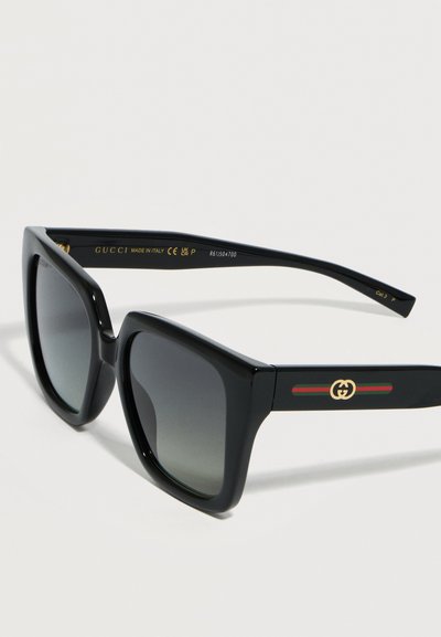 Μαύρα ορθογώνια γυαλιά ηλίου Gucci με σκούρους φακούς διαβάθμισης και χρυσό λογότυπο στις πλευρικές βάσεις που φέρουν πράσινες και κόκκινες ρίγες.