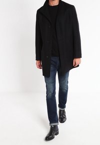 Schwarzer Wollmantel mit breitem Revers und Knopfverschluss, getragen über einem schwarzen Strickoberteil und blauen Jeans. Schlanke schwarze Stiefeletten runden das Outfit ab.