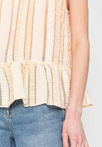Top amarelo claro com riscas texturizadas, barra com rufos e decote em forma de barco. O material parece leve e semi-transparente. Usado com jeans azuis.