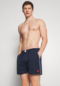 Hombre sin camisa que lleva bañadores azul marino con cordón blanco y logo en la pierna izquierda, de pie contra un fondo claro y liso.