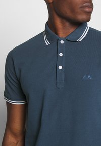 Tmavě modrá polo tričko vyrobené z bavlny, s klasickým límcem s bílými pruhy, čtyřmi knoflíky a malým logem na levé straně.