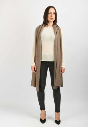 Beige langer Cardigan mit offenem Vorderteil, aus weichem Stoff, getragen über einem leichten Pullover. Schwarze Skinny-Jeans und schwarze, spitz zulaufende High Heels vervollständigen den Look.