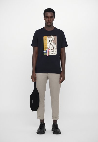 T-shirt en coton bleu marine avec un graphique de lapin de dessin animé et le texte "Paul Smith Since 1970". Modèle portant un pantalon beige clair et des chaussures noires, tenant un sac.