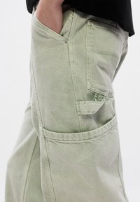 Ανοιχτό πράσινο παντελόνι εργασίας με πολλές τσέπες, υφή υφάσματος και άνετη εφαρμογή, με ορατές ραφές και μεταλλικές λεπτομέρειες.