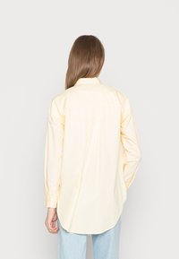 Camisa amarela de botões com mangas longas, vista traseira; possui uma bainha curvada e uma textura suave de algodão. Combinada com jeans azul claro.