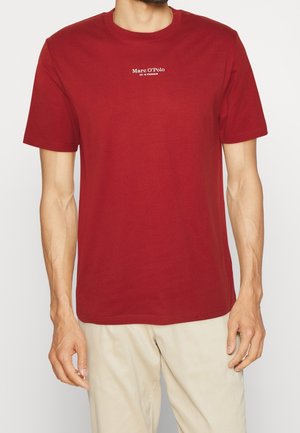 Mann trägt ein schlichtes rotes Marc O'Polo T-Shirt mit kurzen Ärmeln und beigen Hosen vor weißem Hintergrund.