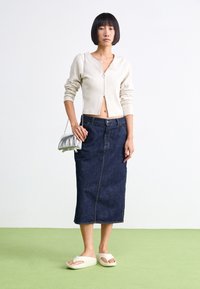 Cardigan beige a costine, gonna a tubino in denim scuro con cucitura frontale, sandali bianchi slip-on e una piccola borsa a tracolla argentata nella mano sinistra.