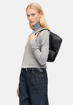 Black leather backpack with a smooth texture, featuring multiple compartments and a sturdy strap. Model wears a light grey turtleneck and dark jeans.