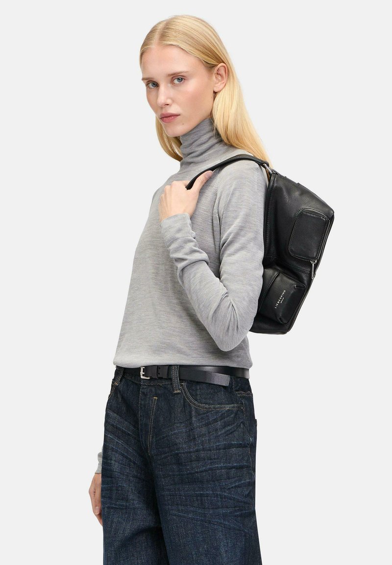 Schwarzer Leder-Rucksack mit glatter Textur, der mehrere Fächer und einen robusten Träger bietet. Das Modell trägt einen hellgrauen Rollkragenpullover und dunkle Jeans.