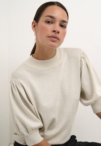 Pulover scurt, cu mâneci scurte, de culoare bej deschis, cu un finisaj texturat, guler ribbat și mâneci ușor plisate. Design minimalist.