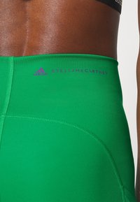 Närbild av en persons midja som bär gröna Adidas x Stella McCartney träningsleggings med logotyper vid midjebandet.