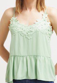 Mentolově zelený top bez rukávů s květinovou krajkovou výšivkou na výstřihu a peplum spodním lemem, vyrobený z lehkého materiálu.