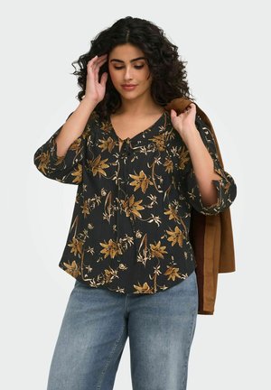 Femme aux cheveux bouclés et foncés, vêtue d'un chemisier noir à fleurs et d'un jean bleu, tenant une veste marron sur son épaule d'une main.