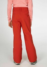 Rode waterdichte snowpants met uitlopende pijpen, elastische tailleband en versterkte naden. De zichtbare combinatie op de schoenen voegt contrast toe.