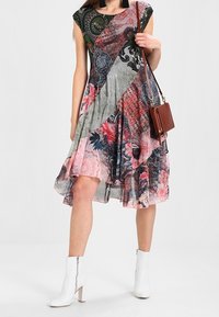 Robe asymétrique à motifs floraux et imprimés dans des tons de rose, gris et vert, associée à des bottines blanches et un sac à bandoulière marron.