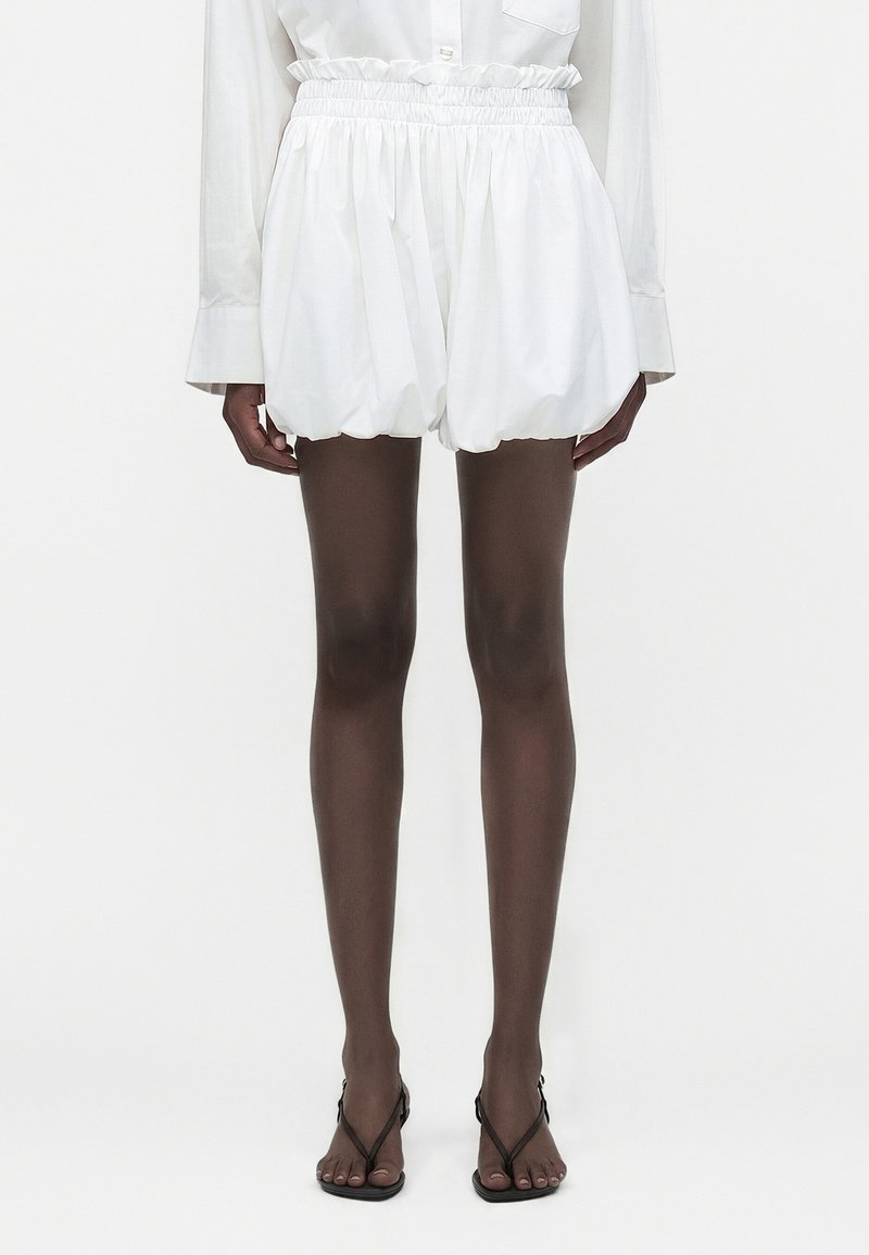 Femme portant un short blanc taille haute froncé, associé à une chemise blanche à manches longues et des sandales noires à brides, debout devant un fond uni.