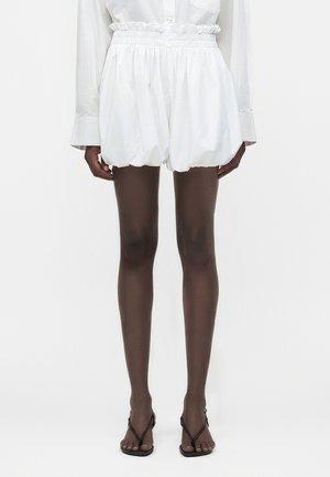 Γυναίκα ντυμένη με λευκά σορτς με ψηλή μέση και ρυτιδωτή λεπτομέρεια, συνδυασμένα με λευκό μακρυμάνικο πουκάμισο και μαύρα σανδάλια με λουριά, στέκεται μπροστά σε μονόχρωμο φόντο.