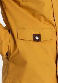 Mosterdgele jas met een gladde textuur, voorzien van een borstzak die is afgewerkt met een bruine stoffen patch en een zilverkleurige drukknoopsluiting.