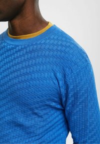 Modrý svetr se vzorem, který má texturovaný design a žlutý kulatý výstřih pod ním. Tkanina připomíná kostkovaný vzor.