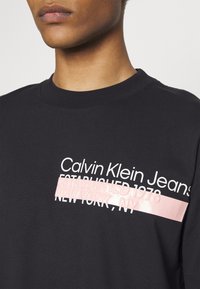 Primer plano de una persona que lleva una camiseta negra con el texto "Calvin Klein Jeans Established 1978 New York, NY" parcialmente cubierto por una franja rosa.