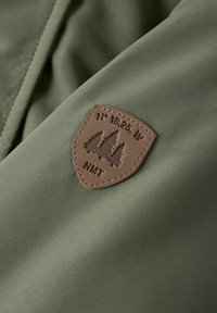 Tessuto verde oliva con una consistenza liscia presenta una toppa a forma di scudo marrone che mostra degli alberi e delle coordinate. La toppa riporta la scritta "N° 18.25 W NMT."