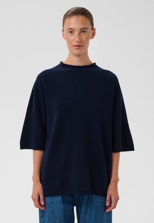 Femme aux cheveux plaqués en arrière portant un pull ample bleu marine foncé avec manches aux coudes et un jean bleu sur un fond clair uni.