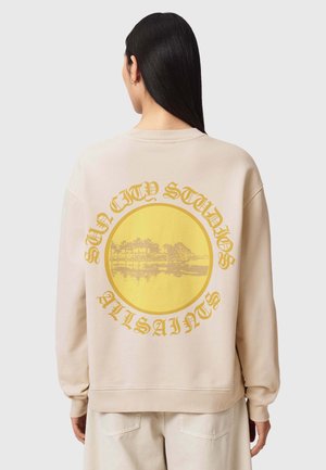 Person trägt beigen Pullover mit gelbem kreisförmigem Motiv von Palmen und dem Schriftzug "Sun City Studios AllSaints" auf der Rückseite.