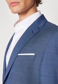 Blauwe textuur blazer met een slim fit, met een geknipt kraag, een wit shirt eronder en een witte pochette in het borstzakje.