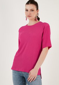 Fuchsia short-sleeve top with a textured finish, round neck, and side slits. Worn with light blue jeans and colorful earrings.