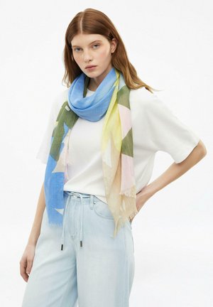 Junge Frau trägt Hellblaue Jeans, weißes T-Shirt und einen mehrfarbigen Schal mit blauen, grünen, gelben und rosa Tönen um den Hals.