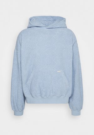 Une personne assise porte un sweat à capuche bleu clair à la texture marquée, un pantalon noir et des bottes noires à lacets, sur un fond uni de studio.