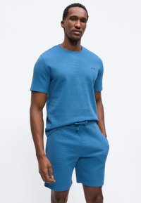 Άνδρας που φοράει κοντομάνικο μπλε T-shirt με υφή και ταιριαστό σορτς με κορδόνι, στέκεται με το ένα χέρι στην τσέπη, μπροστά από απλό φόντο.