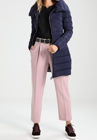 Námořnický péřový kabát se zipem vpředu, kombinovaný se světle růžovými kalhotami vysokého pasu a tmavými teniskami s texturovanými akcenty.