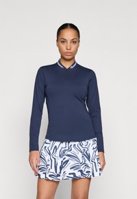 Maglietta a maniche lunghe blu navy con collo a mezza zip e strisce bianche a contrasto, abbinata a una gonna bianca a fantasia con linee astratte blu.