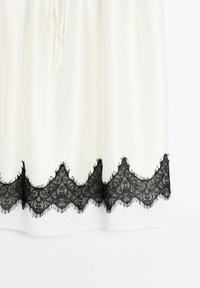 Weißer Stoffrock mit strukturierter schwarzer Spitzenborte am Saum, die ein gewelltes Design und fransige Kanten aufweist. Bänderung in der Taille.